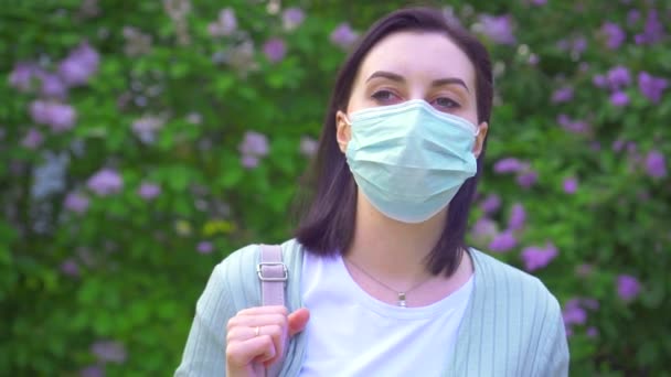 Retrato de uma jovem mulher em uma máscara médica no fundo de plantas com flores no parque — Vídeo de Stock