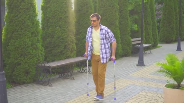 Stylish man with an injury on crutches walking in the Park on a Sunny day — Stock Video