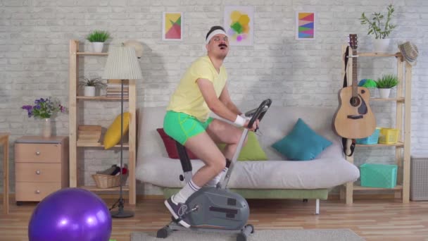 Funny tired athlete from the 80s with a mustache is engaged on a exercise bike in the house slow mo — Stock Video