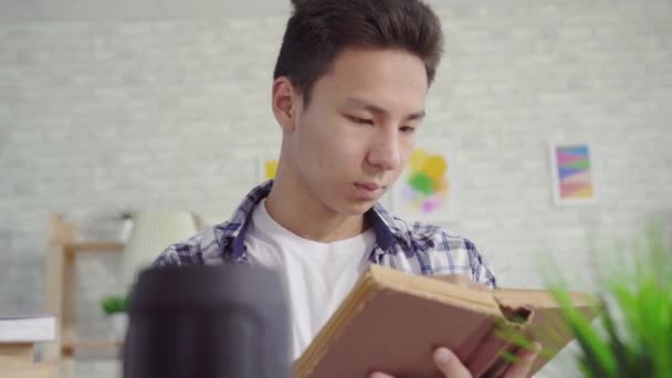 Jovem asiático homem em uma camisa lê um livro e usa um assistente de voz de perto — Vídeo de Stock