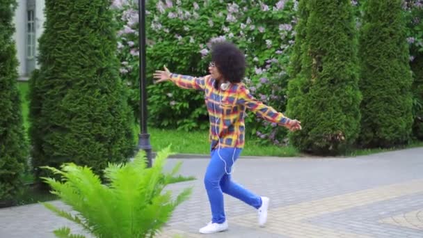 Fröhliche schwarze Frau mit Afro-Frisur und Kopfhörer, die tanzend die Straße hinuntergeht und langsam singt. — Stockvideo