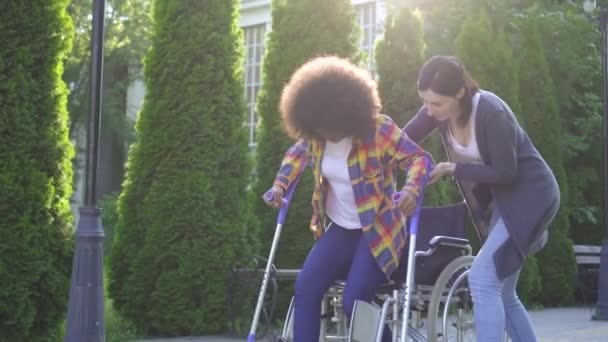 Afrikaanse vrouw met een Afro kapsel uitgeschakeld in een rolstoel in revalidatie leren lopen met de hulp van de assistent — Stockvideo