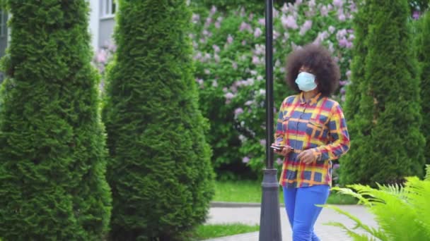 Afro amerikansk kvinna med en afro frisyr i en skyddande medicinsk mask med en smartphone i handen långsam Mo — Stockvideo