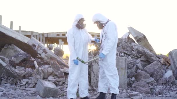 Zwei Wissenschaftler in Schutzanzügen und Masken und messen die Strahlung vor dem Hintergrund der Ruinen bei Sonnenuntergang — Stockvideo
