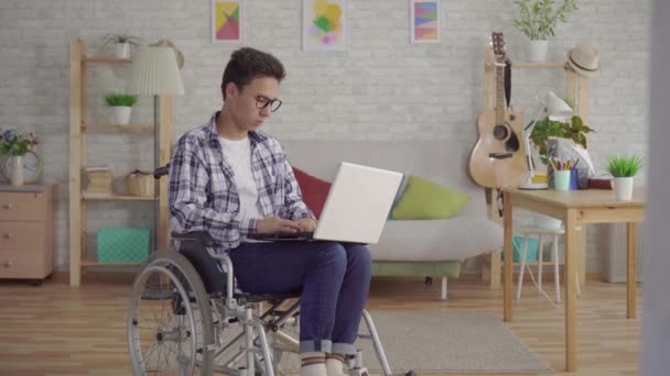 Portret jonge Aziatische man in een stijlvolle bril uitgeschakeld in een rolstoel met een laptop in de woonkamer van het huis — Stockvideo