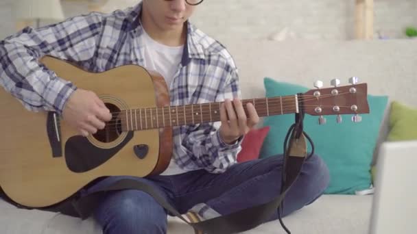 Positiver junger asiatischer Mann mit Hut und stylischer Brille mit Gitarre, der auf dem Sofa im heimischen Wohnzimmer in die Kamera schaut — Stockvideo