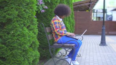 Bir afro saç modeli ile Siyah Amerikalı kadın sokakta bankta oturan bir dizüstü bilgisayar kullanır