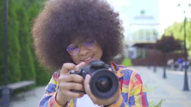 Afrikansk kvinna med en afro frisyr fotograf med en kamera på det urbana landskapet tittar på kameran och leende — Stockvideo