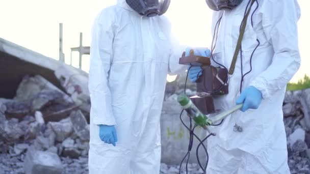 Portrait von zwei Wissenschaftlern in Schutzanzügen und Masken und einem Strahlenüberwacher, die bei Sonnenuntergang Strahlung auf dem Hintergrund der Ruinen einer von Menschen verursachten Katastrophe messen — Stockvideo