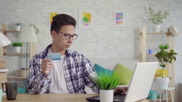 Frustrado jovem asiático homem em uma camisa entra dados com um cartão bancário em um laptop — Vídeo de Stock