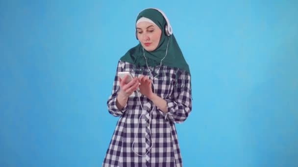 Porträt einer jungen muslimischen Frau mit Kopfhörer und Telefon, die Musik hört und auf blauem Hintergrund tanzt — Stockvideo