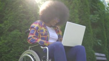 Bir tekerlekli sandalyede engelli bir afro saç modeli ile Smiler afrikalı Amerikalı kadın parkta bir dizüstü sunflare kullanır