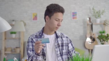 genç bir Asyalı adam bir dizüstü bilgisayarda çalışır ve evin oturma odasında bir Banka kartı tutar ve kameraya bakar