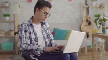 şık gözlüklü genç Asyalı adam tekerlekli sandalyede engelli kişi evin oturma odasında uzaktan dizüstü bilgisayar ile kamera bakarak
