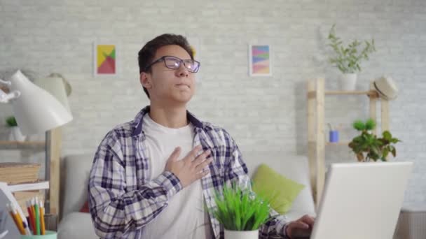 Ung asiatisk man i en skjorta med en hjärtesorg sitter på den bärbara datorn — Stockvideo