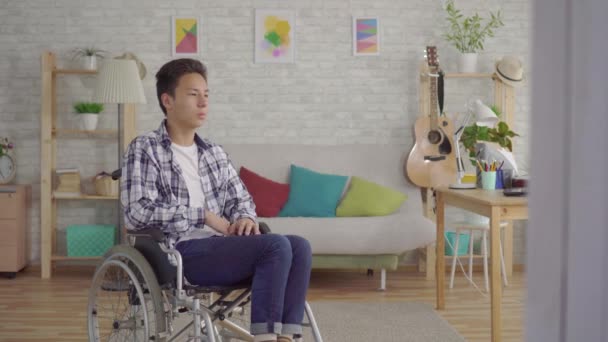 Szomorú fiatal ázsiai férfi fogyatékos a kerekesszékben a nappaliban — Stock videók
