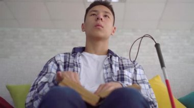 Kör Asyalı genç adam oturma odasında kanepede oturan Braille metin bir kitap okuma yakın