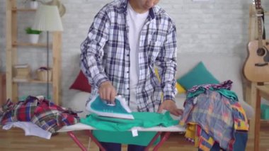 Genç asyalı adam ütü tahtasında elbise ütüleme kameraya bakarak ve gülümseyerek