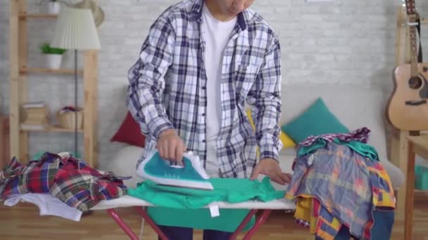 Joven asiático hombre planchando ropa en tabla de planchar mirando a la cámara y sonriendo — Vídeo de stock