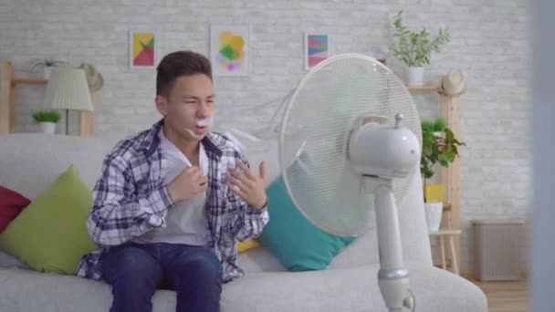 Junger asiatischer Mann sitzt vor einem funktionierenden Ventilator, das Konzept der Hitze — Stockvideo