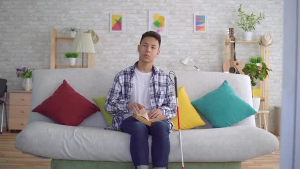 Vak ázsiai fiatalember olvasott könyv Braille-szöveg ül a kanapén a nappaliban — Stock videók