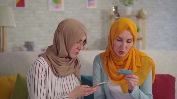 Dua wanita muslim muda yang cantik mengenakan jilbab dengan ponsel dan kartu Bank di tangan mereka berbicara di ruang tamu rumah mereka — Stok Video