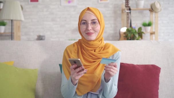 Retrato positivo joven musulmana en hijab sonriente celebración de teléfono inteligente y tarjeta bancaria sentado en el sofá en la sala de estar en casa — Vídeos de Stock