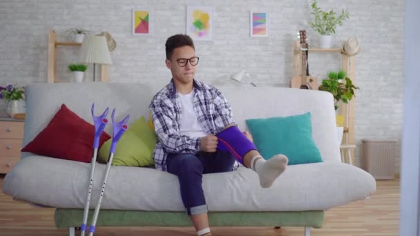 Молодой азиат, сидящий на диване, накладывает упругую повязку на больное колено — стоковое видео