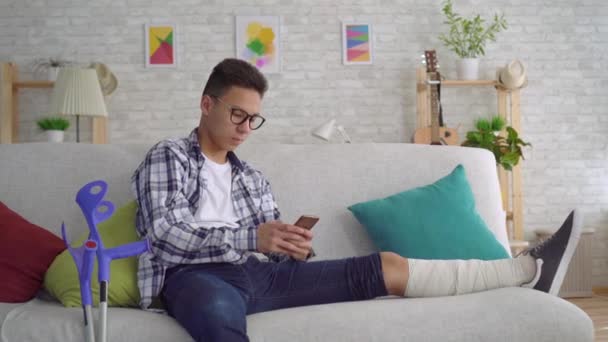 Triste joven asiático con una pierna rota en vendas, acostado en el sofá utiliza el teléfono inteligente y mirando a la cámara — Vídeos de Stock