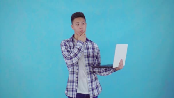 Thoughtful young asian man find solutions with laptop in hand on blue background — Stock Video