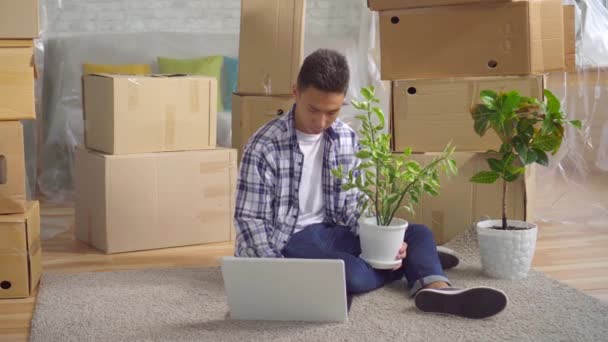 Joven hombre asiático después de mudarse a un nuevo apartamento sentado en un ordenador portátil en el suelo — Vídeos de Stock