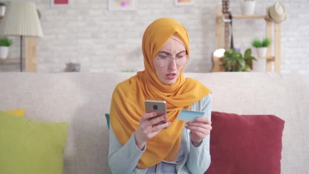 Retrato infeliz e irritado jovem muçulmana no hijab segurando smartphone e cartão bancário sentado no sofá na sala de estar em casa — Vídeo de Stock