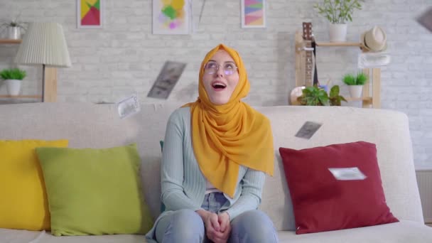 Positive young Muslim woman catches falling banknotes sitting on sofa at home close up — Stock Video