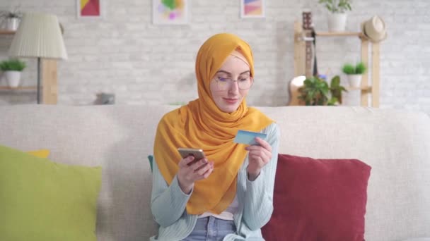 Positivo giovane donna musulmana in hijab sorridente in possesso di smartphone e carta bancaria seduto sul divano in soggiorno a casa — Video Stock