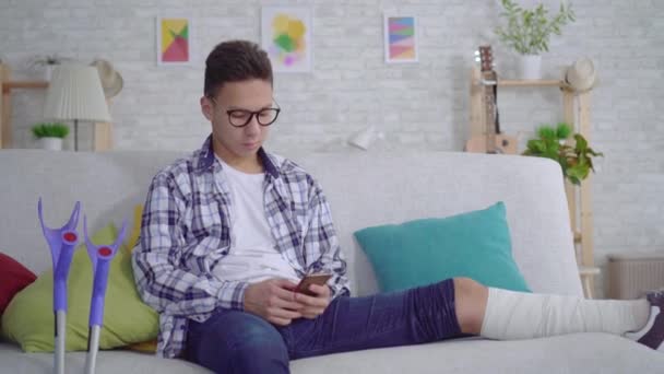 Joven asiático alegre con una pierna rota en vendajes utiliza un teléfono inteligente mirando a la cámara y mostrando los pulgares hacia arriba — Vídeos de Stock