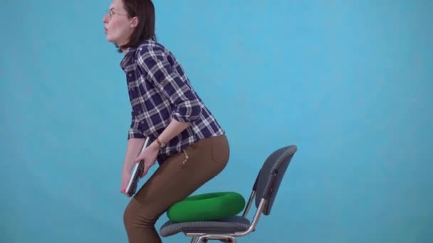 Жінка сидить на ортопедичній подушці на стільці на синьому фоні, концепція геморою — стокове відео