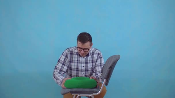 Giovane uomo positivo con occhiali seduto su un cuscino ortopedico da emorroidi su uno sfondo blu — Video Stock