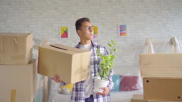 Positive young sian man with a flower and a box in his hand moves to a new apartment — Stock Video