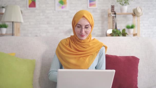 Evde oturma odasında kanepede oturan dizüstü bilgisayar ile pozitif genç Müslüman kadın gülümseyerek kamera bakarak — Stok video