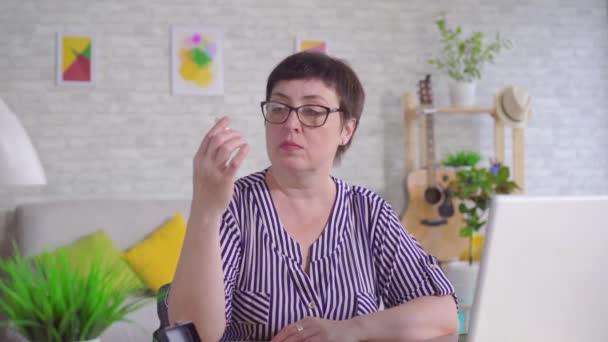 Woman with glasses sitting at a table in the living room holding a hearing aid uses a laptop — Stock Video