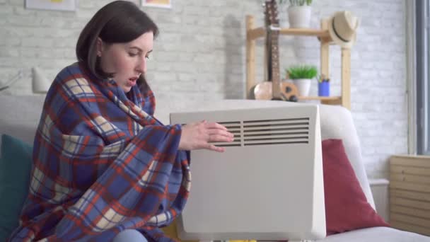 Genç kadın oturma odasında donuyor ve bir elektrikli ısıtıcı yanında kendini ısıtır — Stok video