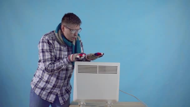 Frozen man covered with frost and gloves heats next to an electric heater on a blue background — Stock Footage Frozen man covered with frost and gloves heats next to an electric heater on a blue background Royalty Free Stock Video