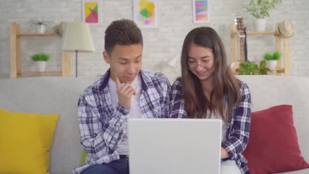 Joven pareja asiática positiva sentada en la sala de estar en el portátil — Vídeos de Stock