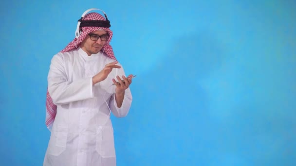 Uomo arabo in cuffia ascoltare musica in piedi su uno sfondo blu — Video Stock