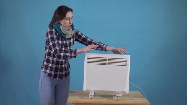 Mujer congelada en la bufanda siguiente se calienta con un calentador eléctrico sobre un fondo azul — Vídeos de Stock