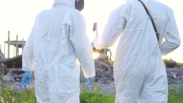 Retrato dos científicos en trajes de protección y máscaras y un dosímetro, caminar medir la radiación en el fondo de las ruinas de un desastre causado por el hombre de cerca al atardecer — Vídeo de stock