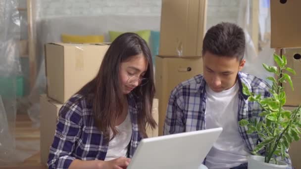 Junges asiatisches Paar fährt in eine neue Wohnung, sitzt auf dem Boden und benutzt einen Laptop aus nächster Nähe — Stockvideo