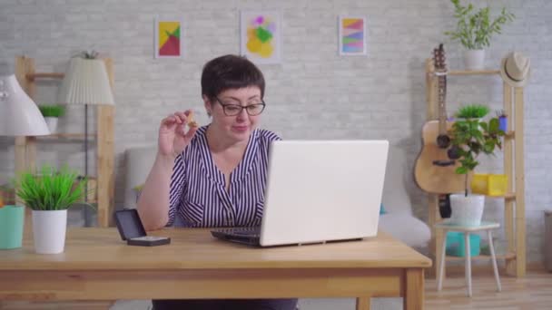Улыбающаяся женщина в очках, сидящая за столом в гостиной с слуховым аппаратом, использует ноутбук — стоковое видео