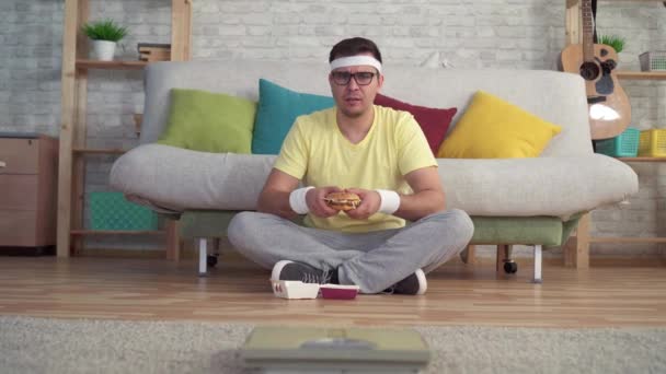 Vicces sportos férfi sportruházat ül a padlón nézi a fürdőszoba mérlegek eszik egy Burger — Stock videók