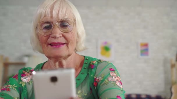 Anciana con el pelo gris utiliza un teléfono inteligente sentado en una mesa en la sala de estar de cerca — Vídeo de stock